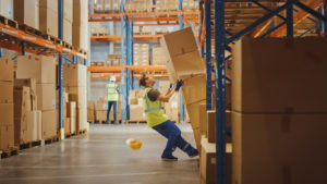 Warehouse Worker Has Work Related Accident. He is Falling Down BeforeTrying to Pick Up Heavy Cardboard Box from the Shelf