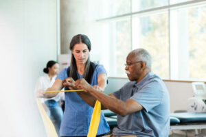 The mid adult female physical therapist teaches the senior adult man how to exercise with the elastic band.