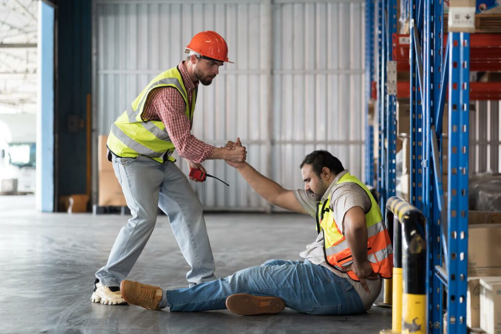 A male warehouse worker is assisting a colleague who had an accident due to a work injury while in the warehouse.